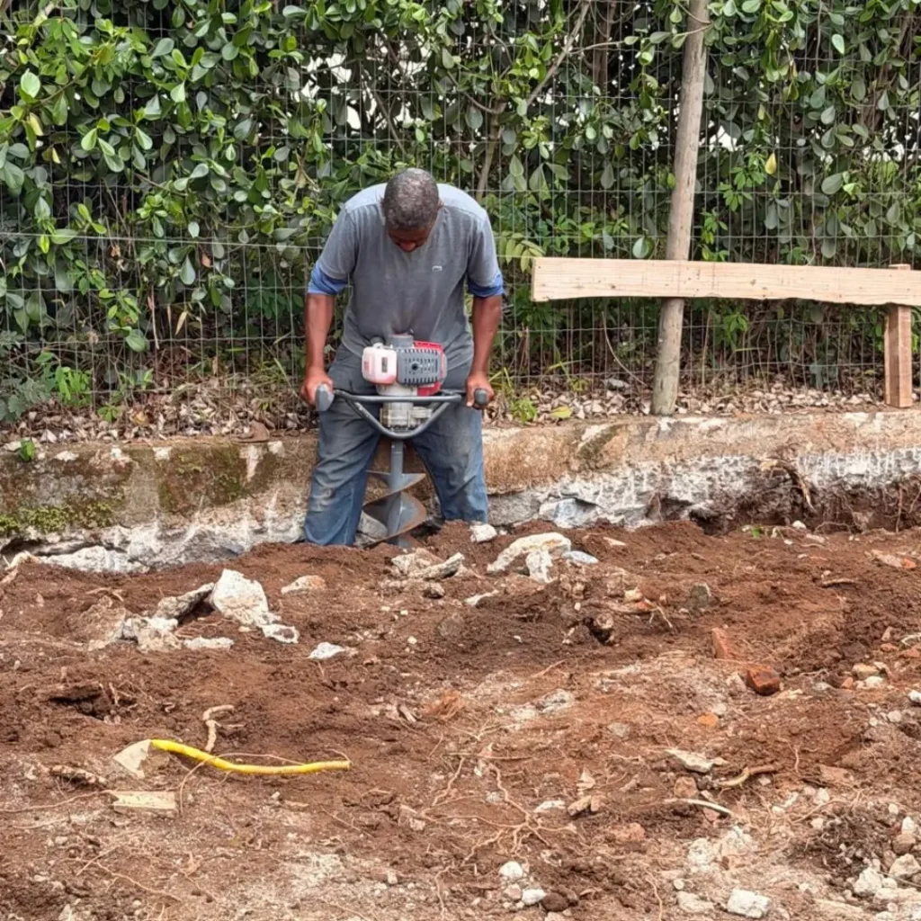 Operário utilizando perfurador de solo mecânico para preparação de alicerce, garantindo agilidade e fim do esforço manual na obra.